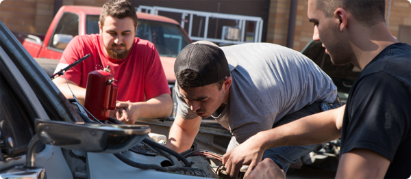 Three automotive students inspect the engine of a car to asses necessary repairs.
